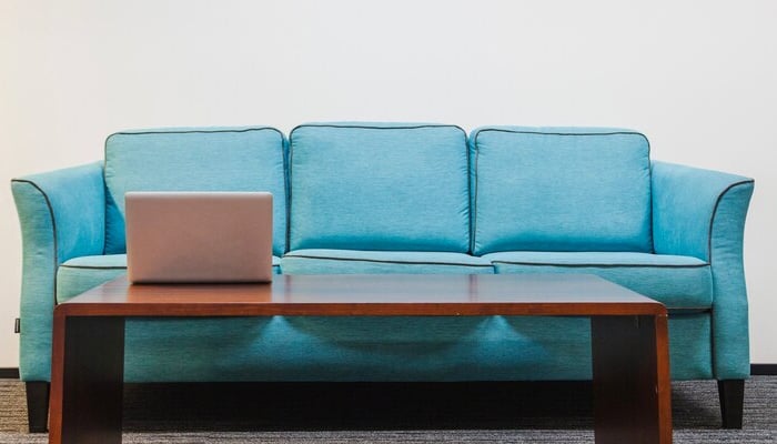 A minimalist living room setup featuring a bright turquoise three-seater sofa and a simple rectangular wooden coffee table. A silver laptop sits on the table, and the background includes a white wall and a gray carpeted floor, creating a clean and modern aesthetic.