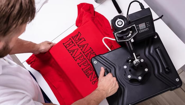 A man using a heat press machine to print the phrase "MAKE IT HAPPEN!" in bold black letters on a red T-shirt.