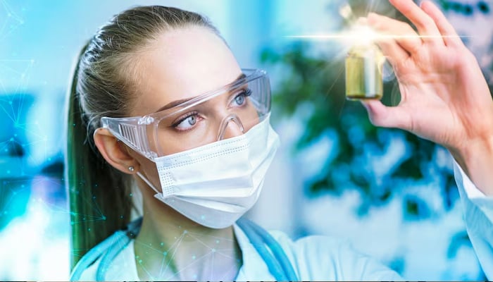 A scientist wearing protective gear, including safety goggles and a mask, closely examines a vial under a bright light in a laboratory setting.