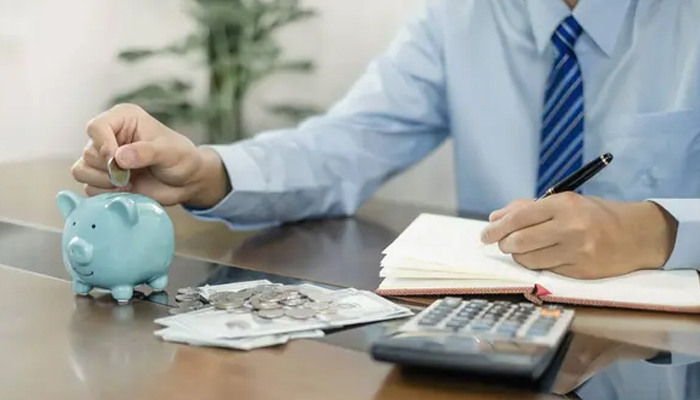 A person placing a coin into a piggy bank, surrounded by cash, coins, a notebook, and a calculator, symbolizing financial management and savings.