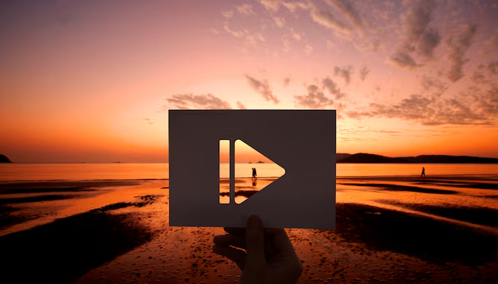 A hand holds a paper cutout of a play button against a vibrant sunset over a beach, with two people walking in the distance on the wet sand reflecting the colorful sky.